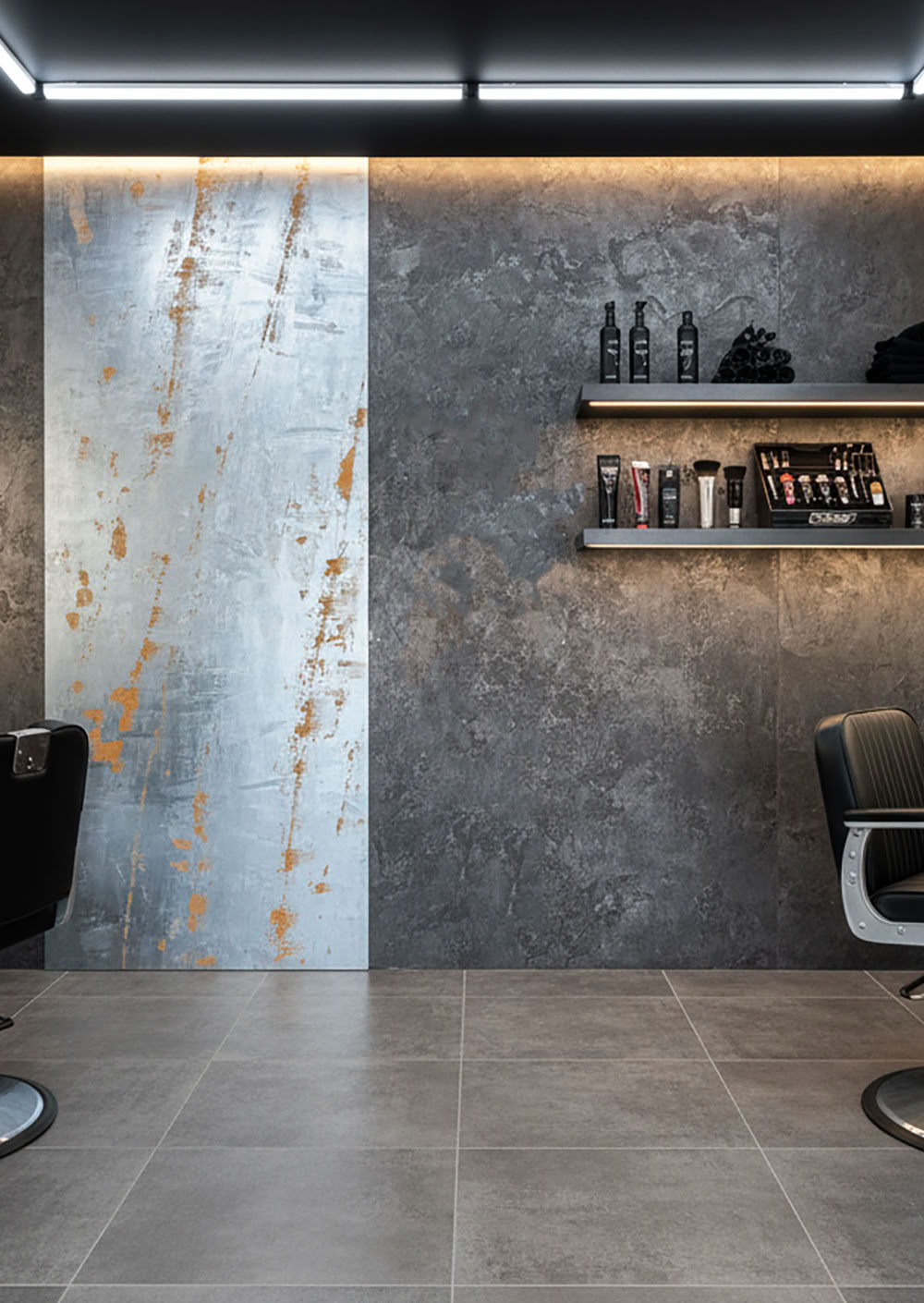Moderner Friseursalon mit zwei Friseurstühlen, Spiegeln an den Wänden, Regalen mit Pflegeprodukten und einer metallischen Wandverkleidung.