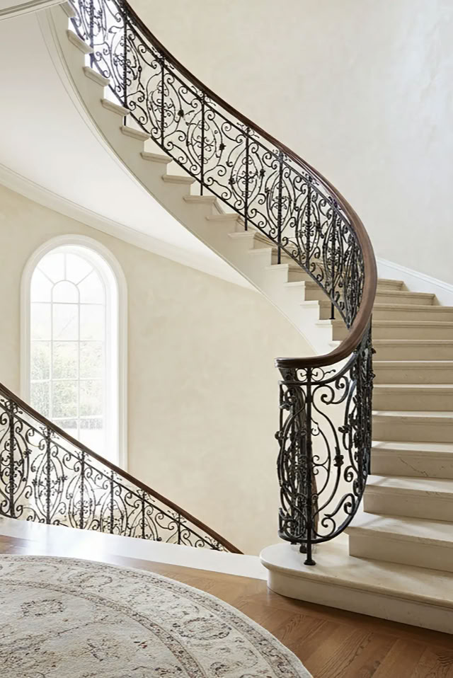 glaMUR Waende in W101 Elfenbein. Geschwungene Treppe mit kunstvollem Geländer, drei hohe Fenster im Hintergrund und ein runder Teppich auf dem Holzboden.