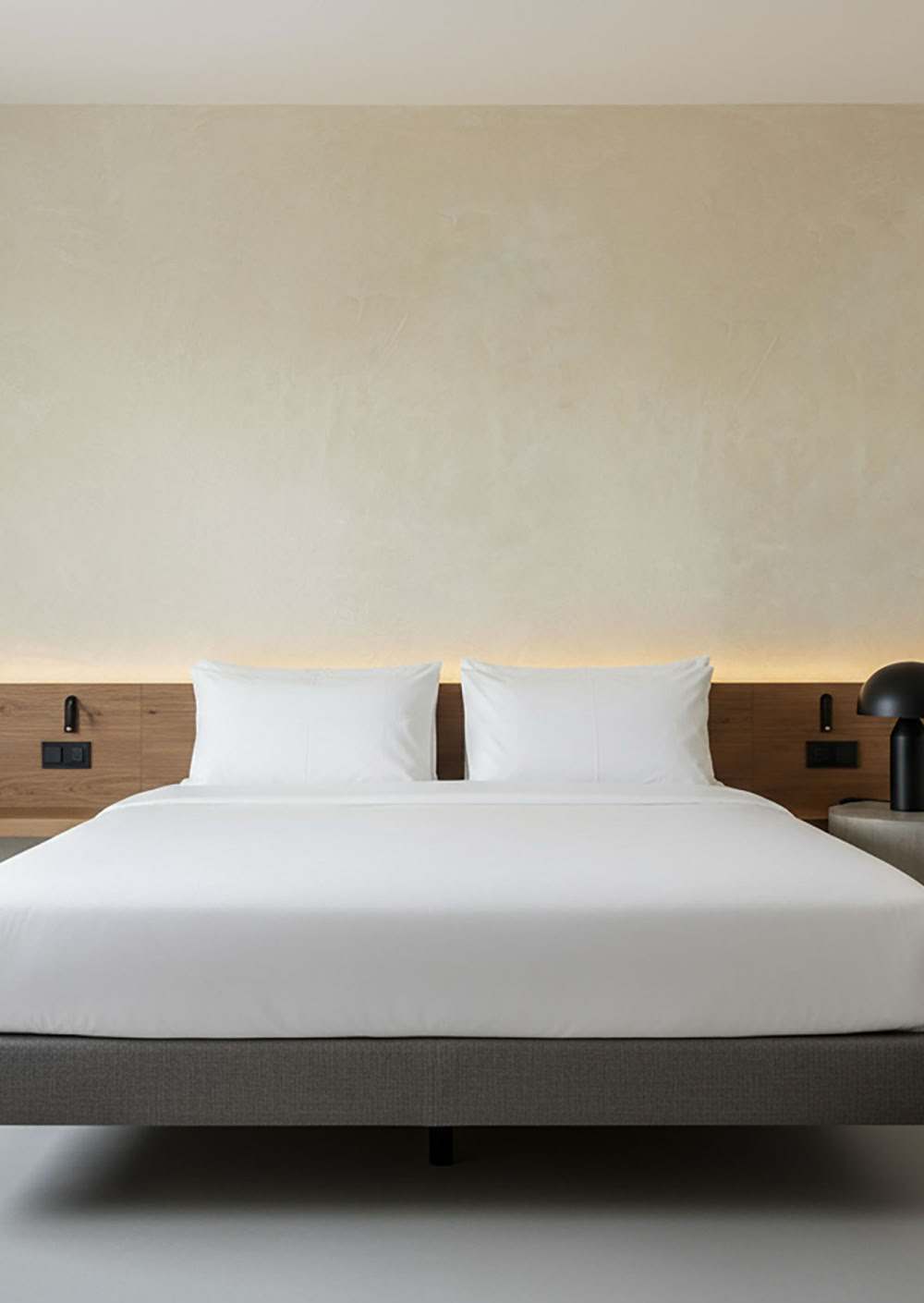 Modernes Hotelzimmer mit einem großen Bett, weißen Bettlaken, zwei Nachttischen aus Holz, einer Wand mit Paterra Farbe Elfenbein